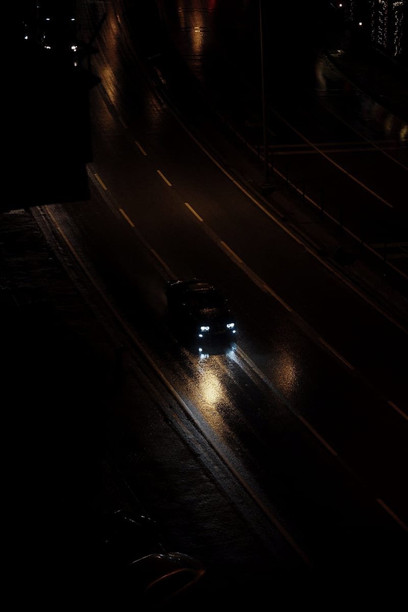 A single car on a wet road at night, headlights streaking across the soaked asphalt